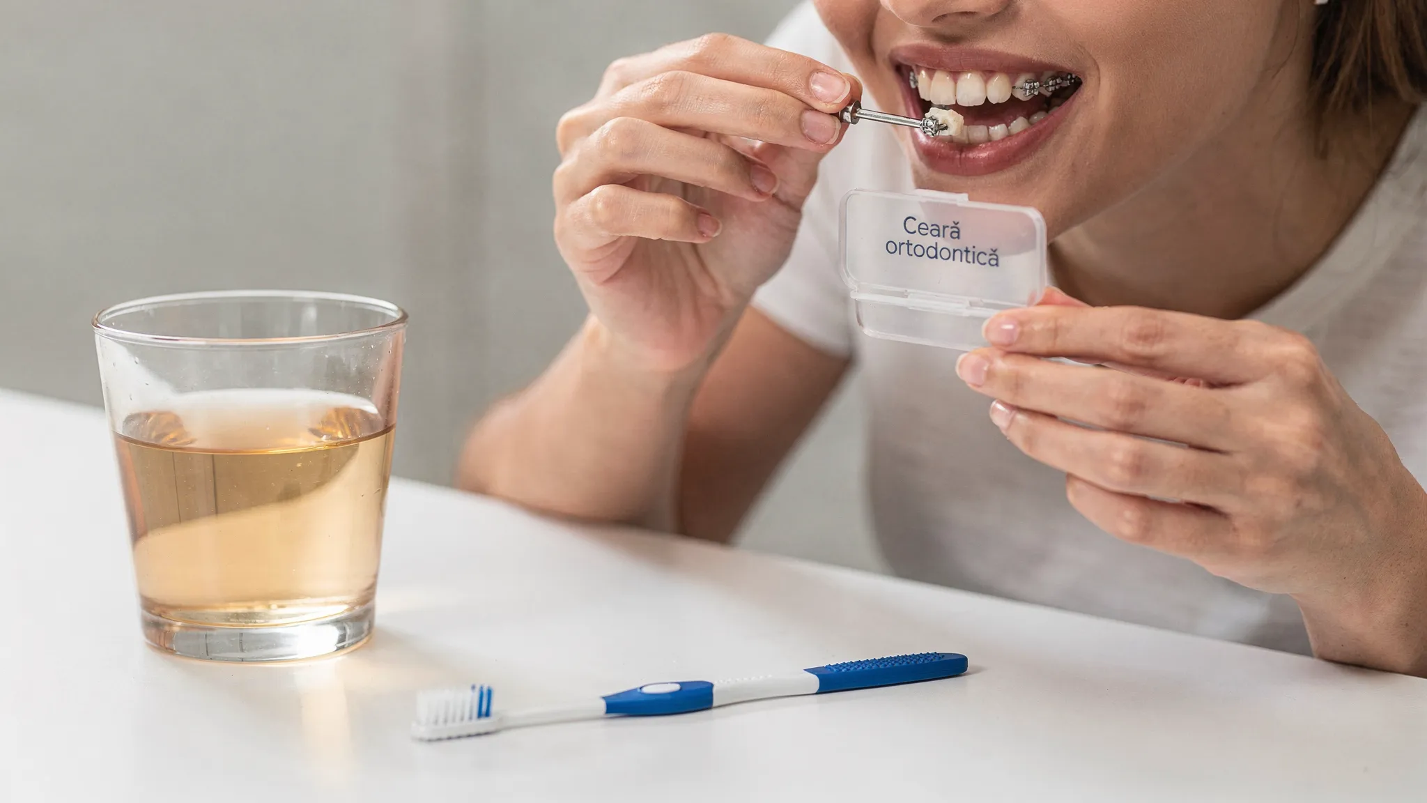 O persoană ținând o cutie mică de ceară ortodontică și aplicând o bucățică pe un bracket, cu un pahar cu apă sărată și o periuță interdentară pe masă.