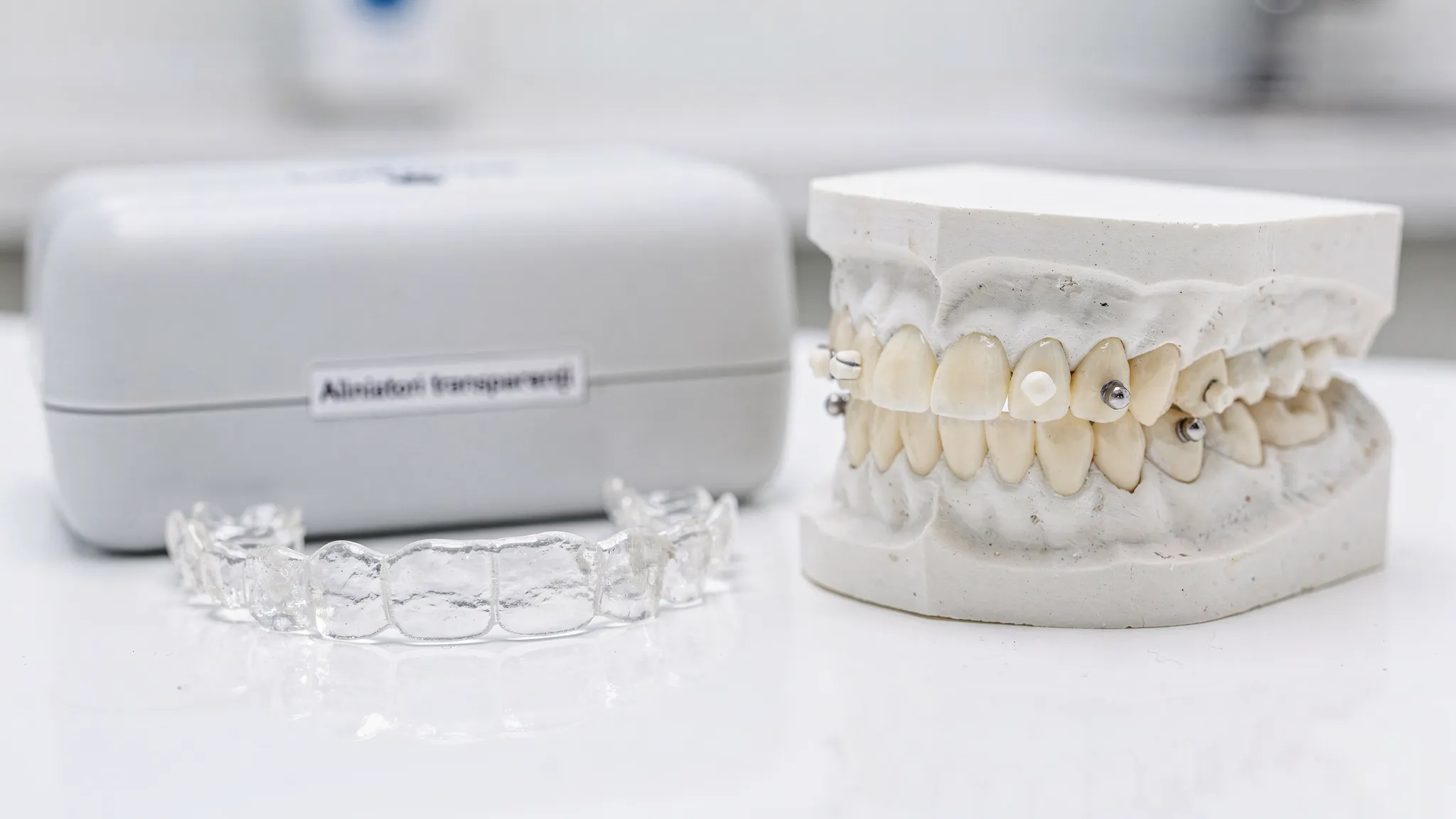 Set de alignere transparente lângă o cutie de depozitare și un model dentar cu mici atașamente vizibile pe câțiva dinți, imagine clinică luminoasă.