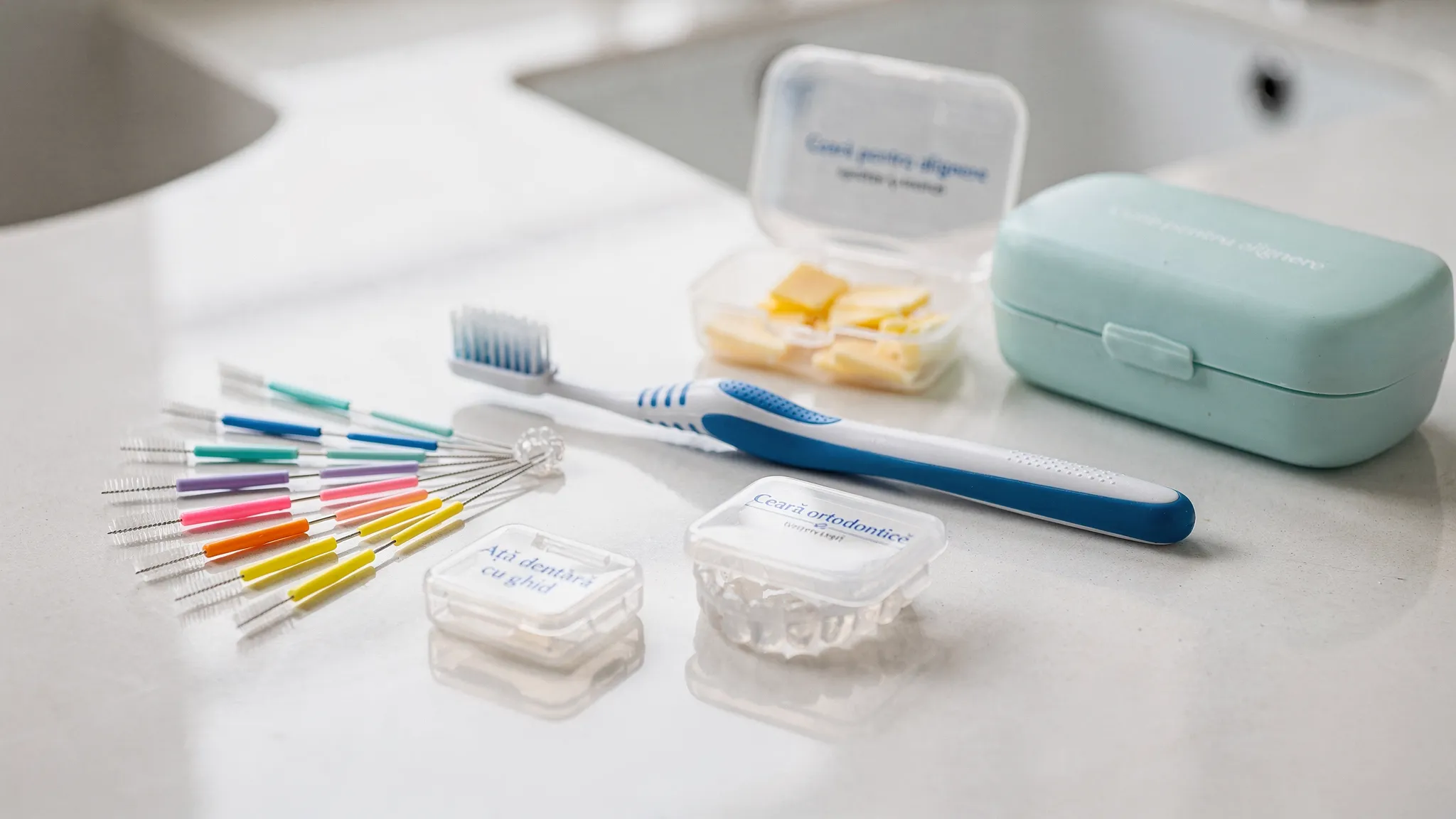 Set de îngrijire pentru aparat dentar: periuță ortodontică, periuțe interdentare, ață dentară cu ghid, ceară ortodontică și o cutie pentru alignere, fotografiate pe o suprafață curată.