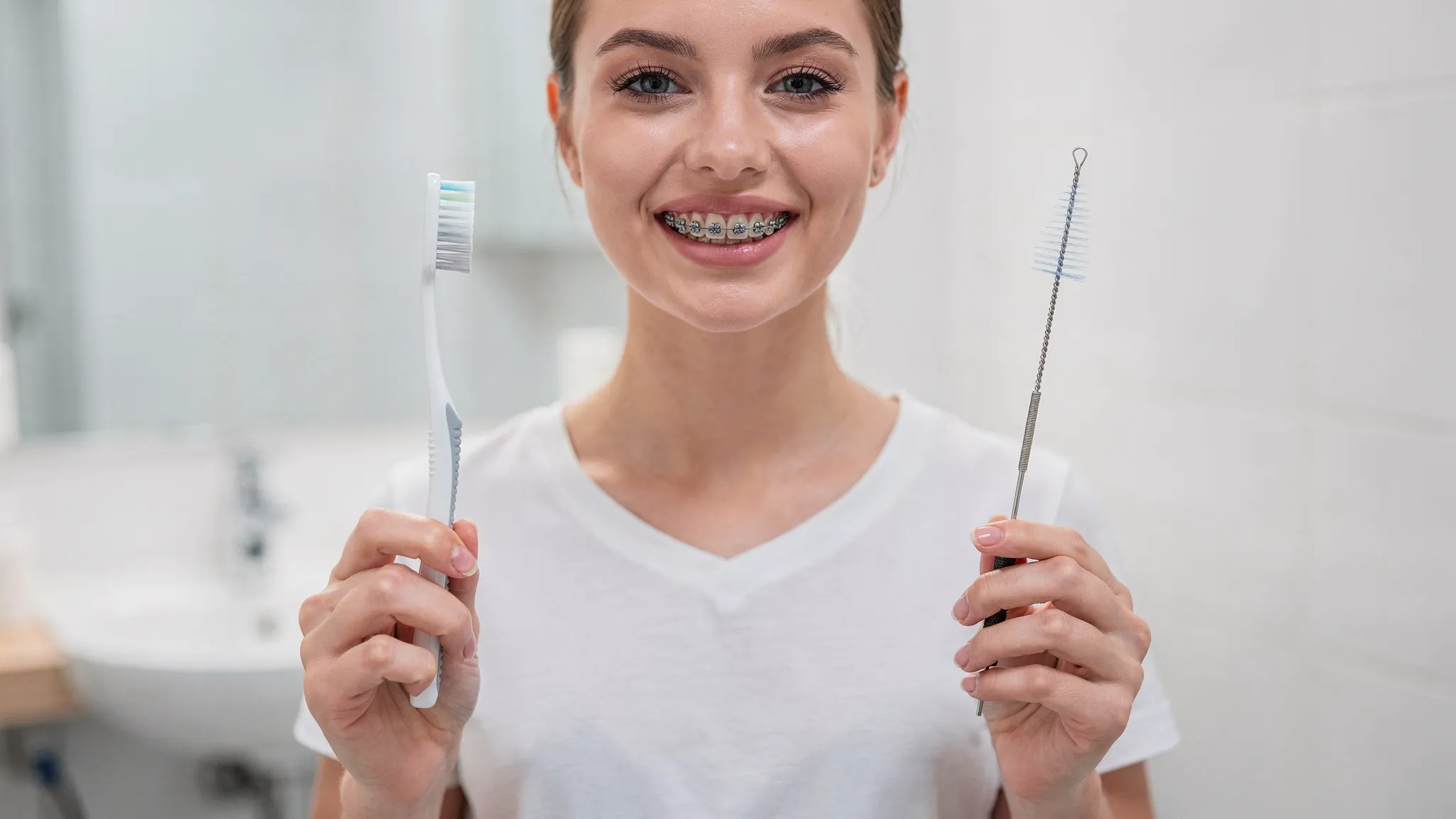 Persoană în baie, ținând o periuță de dinți și o periuță interdentară, în timp ce se vede un aparat dentar fix pe dinți; imagine educativă despre igiena corectă cu brackeți.