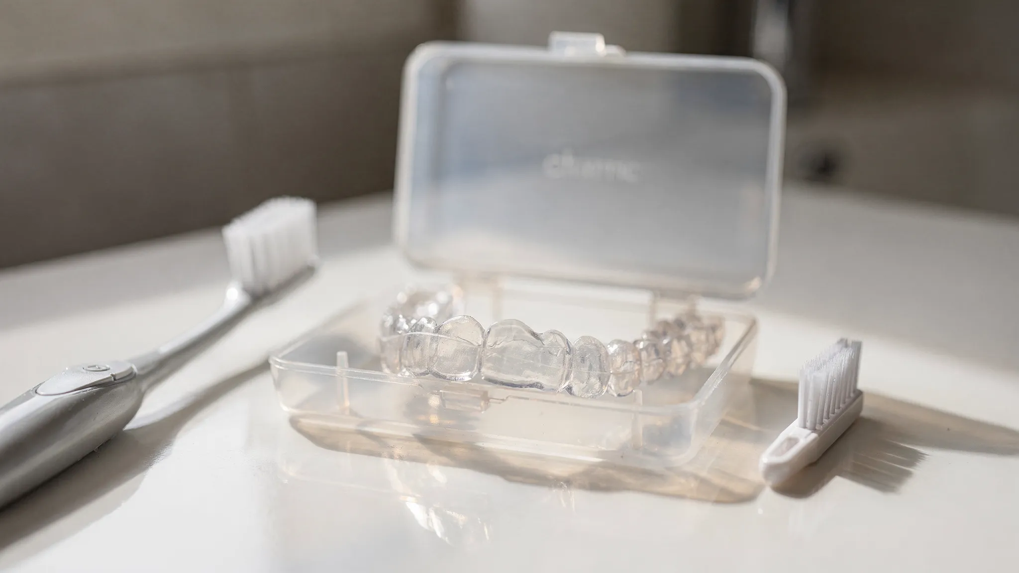 Set de alignere transparente într-o cutie, lângă o periuță de dinți și o periuță mică pentru curățare; scenă de igienă pentru alignere, fără ecrane sau texte vizibile.