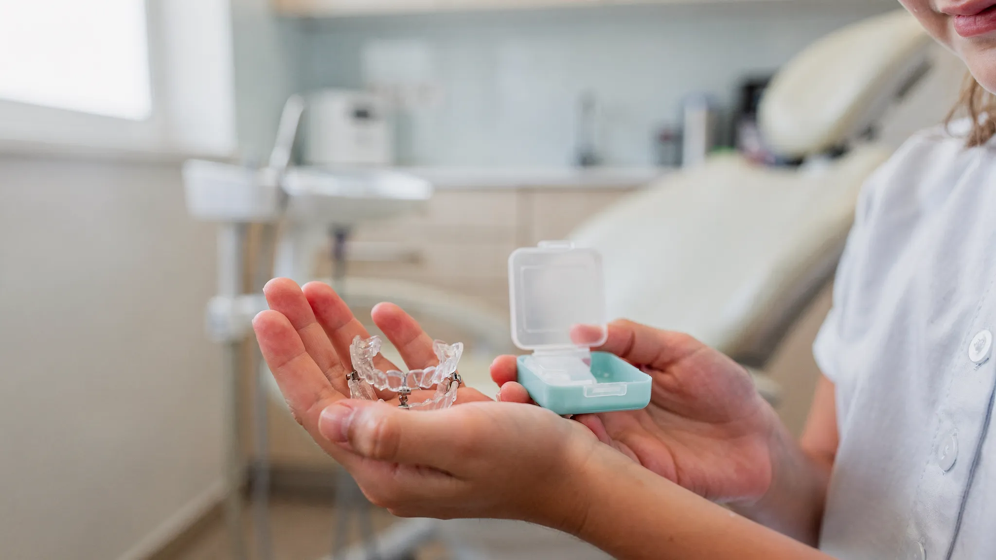 Un copil ține în palmă un aparat ortodontic mobil și o cutie de depozitare; pe fundal se vede, ușor defocalizat, un cabinet stomatologic luminos.