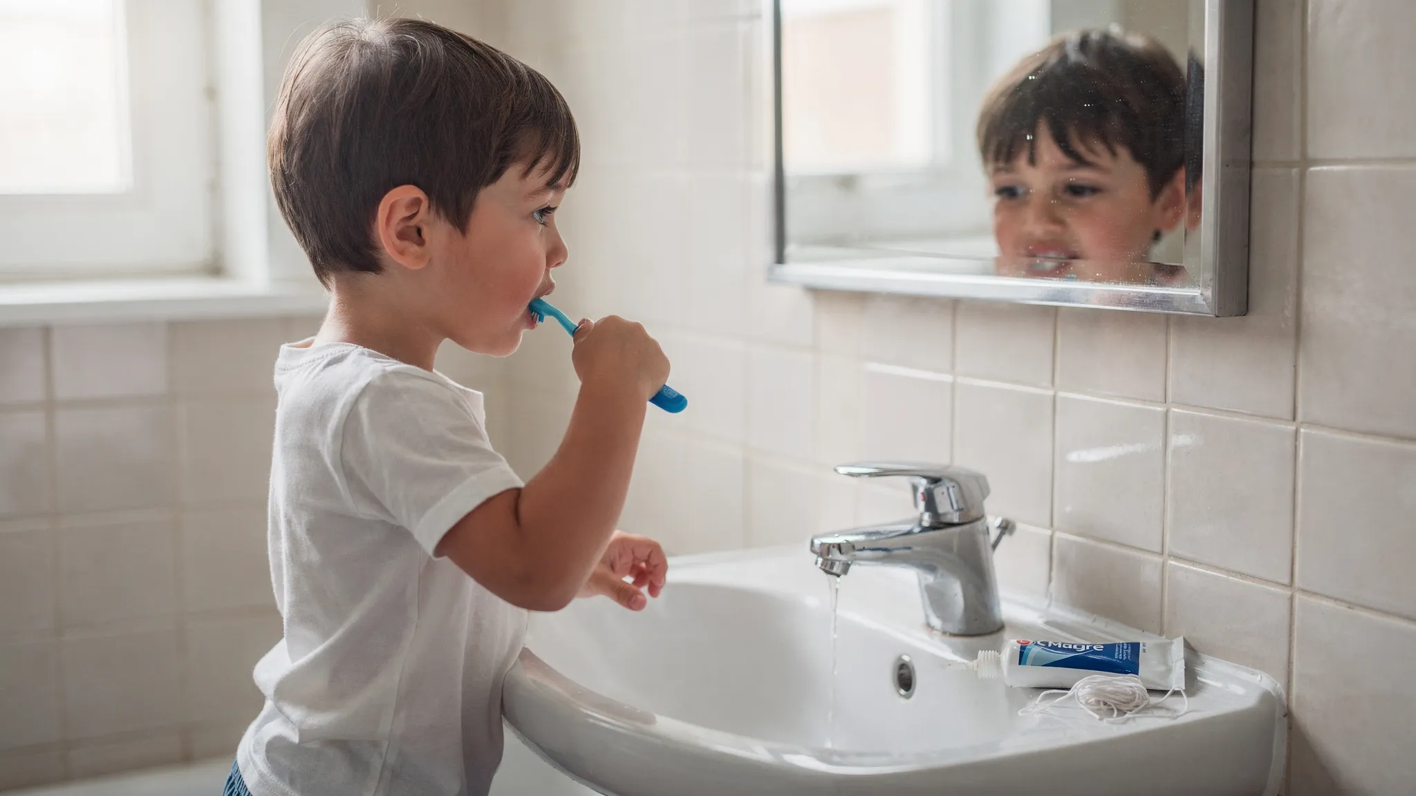 Copil în baie, ținând o periuță de dinți și periaj atent; pe chiuvetă se văd o pastă de dinți și o ață dentară; atmosferă luminoasă, fără ecrane sau texte.