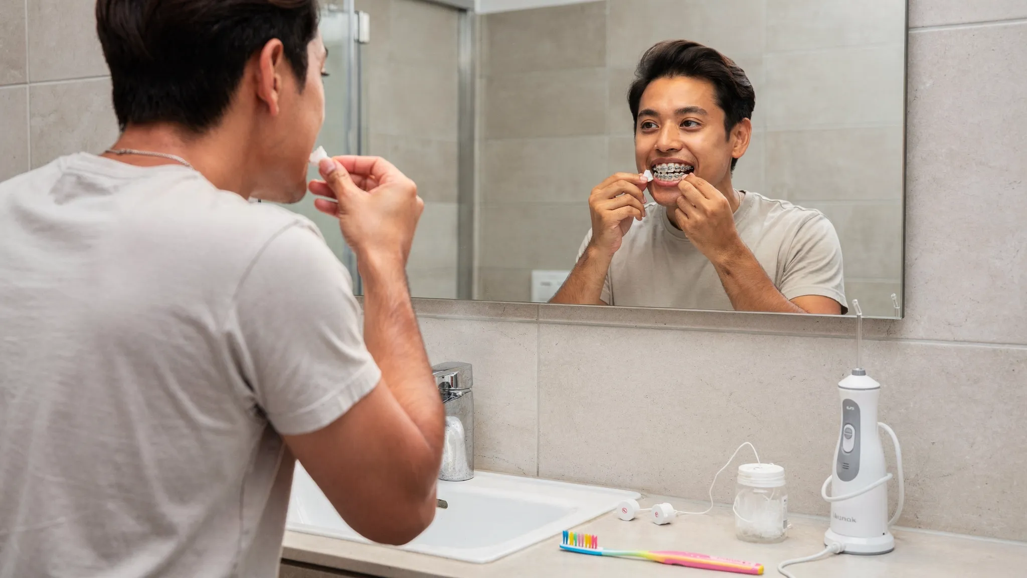 O persoană în baie, în fața oglinzii, arătând cum aplică ceară ortodontică pe un bracket; pe blat se văd o periuță de dinți, ață dentară și un duș bucal.