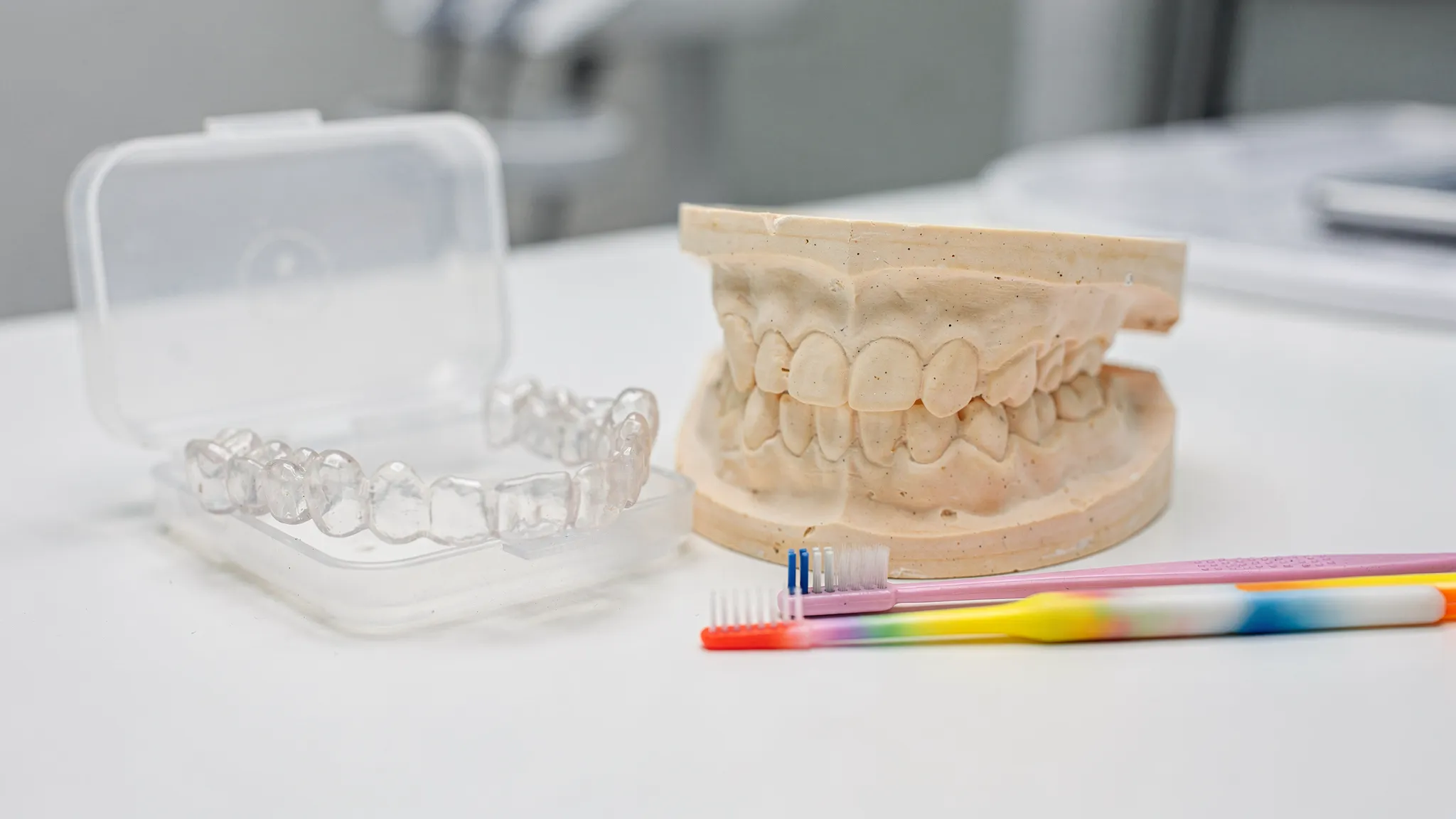 Retainer transparent și model dentar pe o masă de consultație ortodontică, alături de o periuță interdentară și o cutie de protecție pentru gutieră.