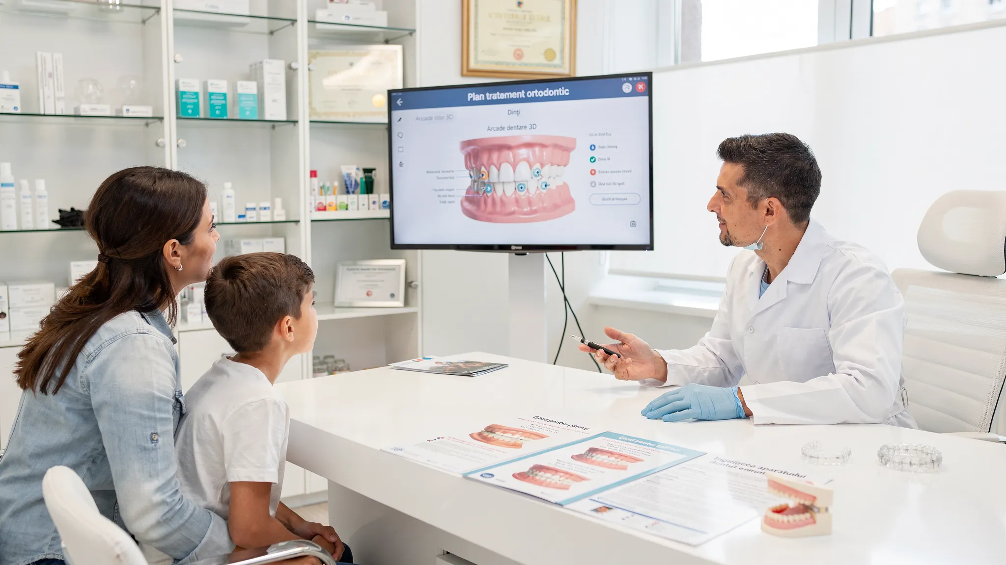 Scenă într-un cabinet modern de ortodonție: medicul arată unui părinte și unui copil un model 3D al arcadelor dentare pe un ecran orientat corect, cu discuție calmă și materiale educative pe birou.