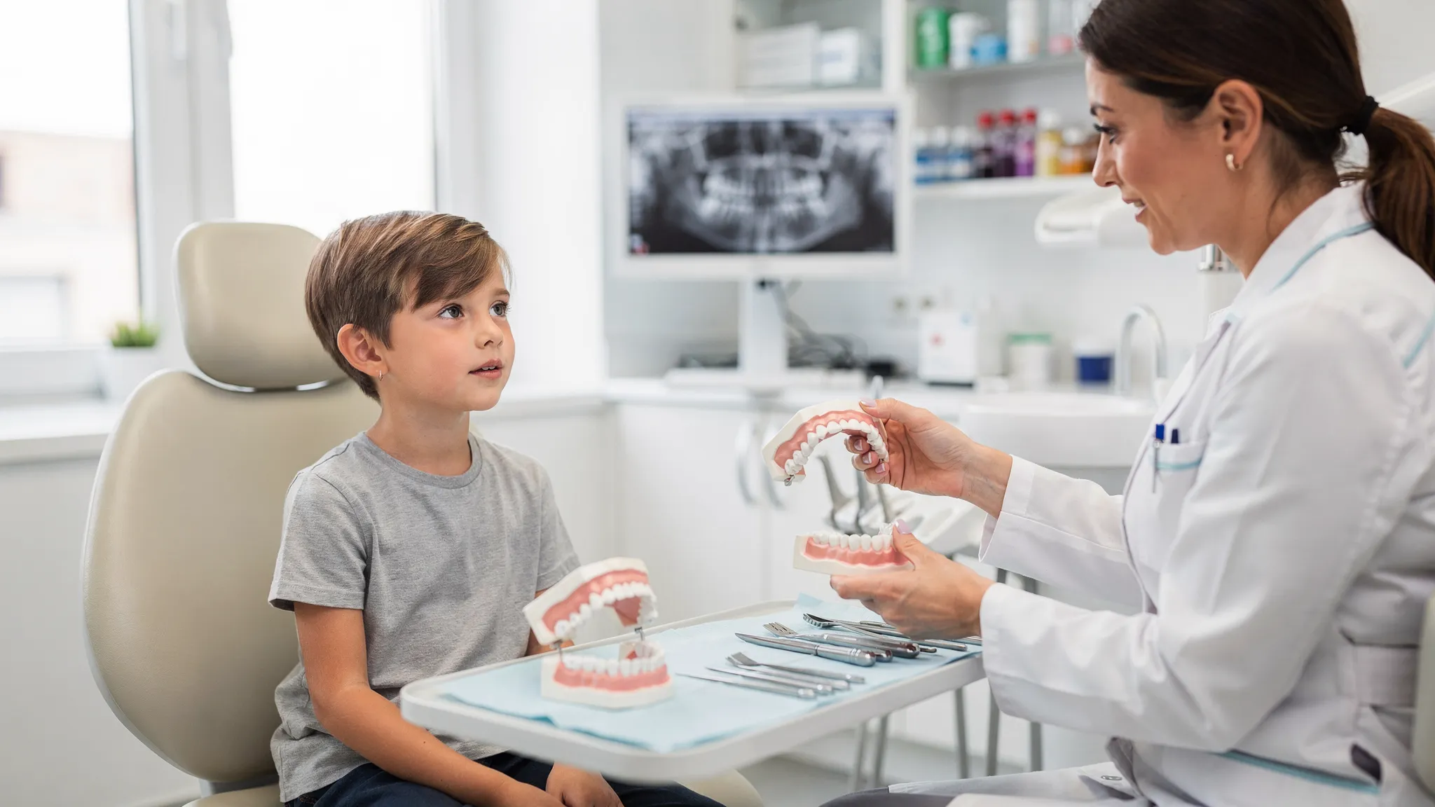 Un copil la consultație ortodontică, cu medicul explicând pe un model de arcadă dentară diferența dintre maxilar îngust și arcadă normală, într-un cabinet luminos.