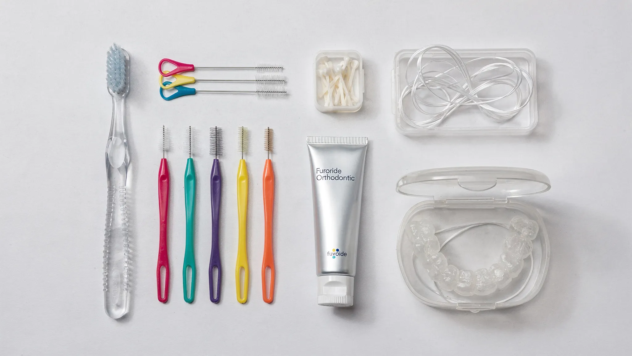 Kit de igienă pentru aparat dentar cu periuță ortodontică, periuțe interdentare, ață dentară specială, ceară ortodontică, pastă cu fluor și cutie pentru alignere transparente așezate ordonat pe o suprafață curată.