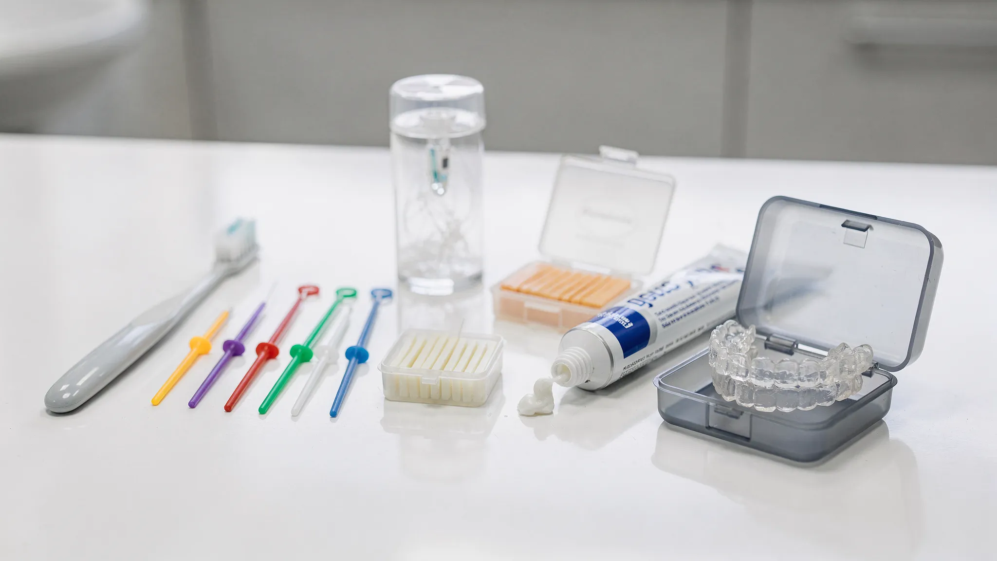 Produse de îngrijire ortodontică așezate ordonat pe o suprafață curată: periuță cu peri moi, periuțe interdentare, ață dentară ortodontică, ceară ortodontică, pastă cu fluor și cutie pentru alignere transparente.