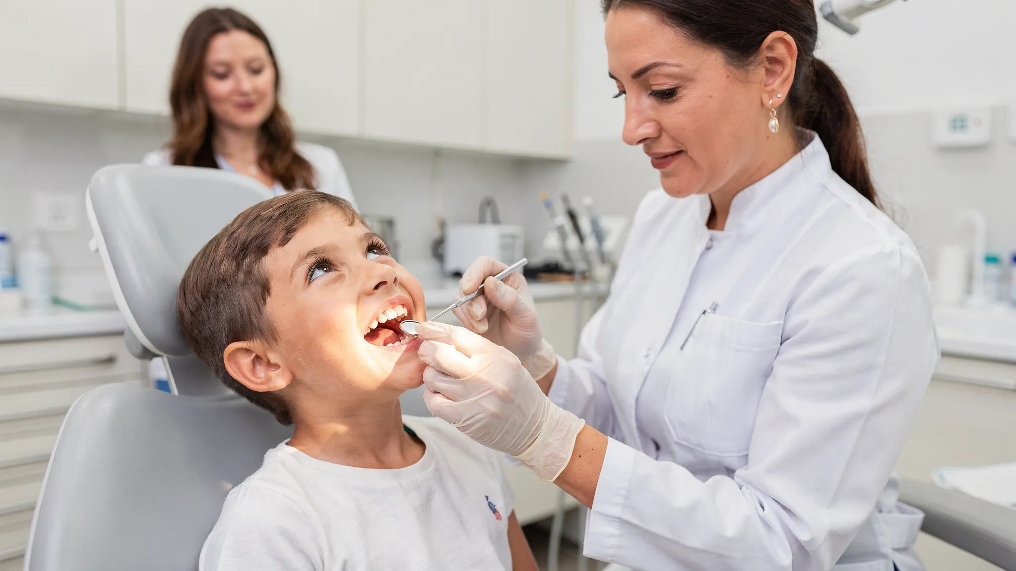 Un copil zâmbește într-un cabinet ortodontic, iar medicul îi evaluează mușcătura și poziția dinților cu ajutorul unei oglinzi dentare. Pe fundal se află un părinte atent și un spațiu clinic luminos.