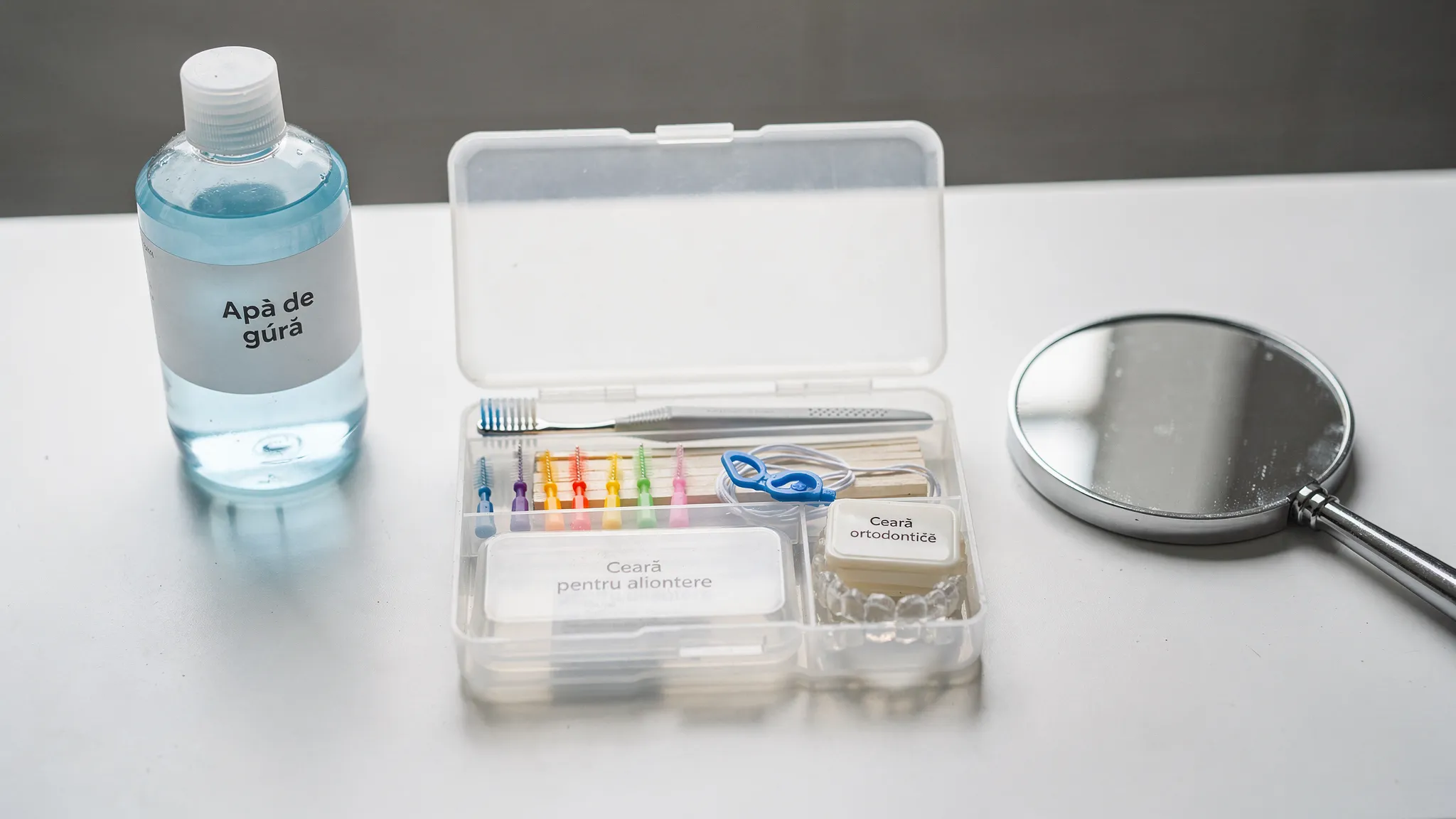 Un kit de ortodonție pentru acasă așezat pe o suprafață curată, cu periuță ortodontică, periuțe interdentare, ață dentară cu dispozitiv de trecere, ceară ortodontică, cutie pentru alignere, apă de gură și o oglindă mică.