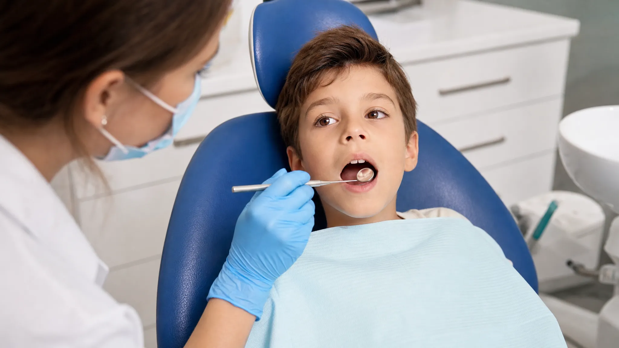 Un copil stă pe scaunul stomatologic în timp ce medicul examinează atent dinții frontali cu o oglindă dentară pentru a observa mici pete albe pe smalț.