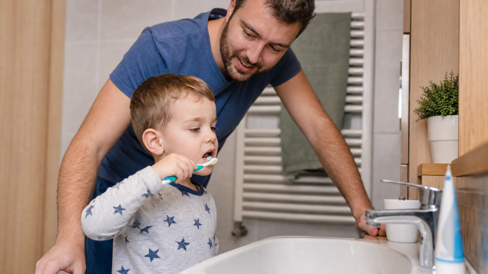 Un părinte supraveghează un copil mic care se spală pe dinți în baie, cu o cantitate mică de pastă cu fluor pe periuță, lângă chiuvetă.